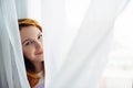 Cute young red-haired girl looks out from the white curtain, close-up, copy space. Portrait of beautiful smiling woman with Royalty Free Stock Photo