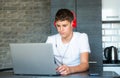 Cute young teenager in white shirt sitting behind desk in kitchen next to laptop and study. Serious boy in earphones Royalty Free Stock Photo