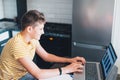 Cute young teenager in white shirt sitting behind desk in kitchen next to laptop and study. Serious boy in earphones Royalty Free Stock Photo