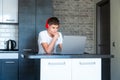 Cute young teenager in white shirt sitting behind desk in kitchen next to laptop and study. Serious boy in earphones Royalty Free Stock Photo