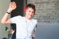 Cute young teenager in white shirt sitting behind desk in kitchen next to laptop and study. Serious boy in earphones Royalty Free Stock Photo