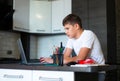 A cute young teenager in a white t-shirt sitting behind a desk in a kitchen next to a laptop and study. Serious boy makes homework Royalty Free Stock Photo