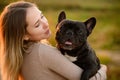 Cute young woman holding French bulldog, smiling at sunset while walking. Favorite dog in the arms of the owner in the Royalty Free Stock Photo