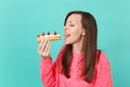 Cute young woman in knitted pink sweater with closed eyes holding in hand, eating eclair cake isolated on blue turquoise