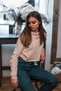 Cute young woman in a stylish pink sweatshirt in fashionable blue jeans is resting sitting on a chair in a cozy bedroom near a Royalty Free Stock Photo