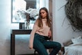 Cute young woman in a stylish pink sweatshirt in fashionable blue jeans is resting sitting on a chair in a cozy bedroom near a Royalty Free Stock Photo