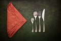 A cutlery set and terracotta linen napkin on a dark green background. Spoons, fork and knife, flat lay