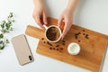 Cutting board with cup of fresh coffee, milk, coffee beans, phone, female hands and plant branch on white background, top view Royalty Free Stock Photo