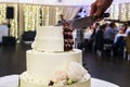 Cutting The Multitiered White Decorated Wedding Cake In A Banquet Hall Background. Wedding Cake Is Cut With Knife. Cake Ornament.