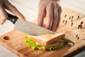 Cutting sandwich with bread, cheese, salad and ham with hands on wooden cutting board with knife Royalty Free Stock Photo