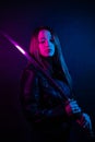 Cyber girl in a black leather jacket at dusk holds a katana. A woman in a club with a colored pink-blue light holds an Asian sword Royalty Free Stock Photo