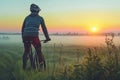 cyclist with bike watching a sunrise over a misty meadow