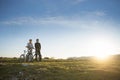 Cyclist couple with mountain bikes standing on the hill under the evening sky and enjoying bright sun at the sunset.