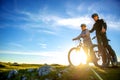 Cyclist couple with mountain bikes standing on the hill under the evening sky and enjoying bright sun at the sunset.