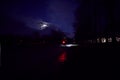 A cyclist with powerful lights and safety vest rides in a dark urban park under a cloudy full moon at dusk