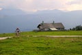 A cyclist on the road between the fields with view on the mountains and a house in the clouds.