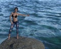 Illustration of a beautiful woman wearing a bikini top and jeans standing on a boulder surrounded by water with a hand and thumb Royalty Free Stock Photo