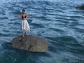 Illustration of a beautiful woman wearing a bikini top and a white skirt standing on a boulder surrounded by water with hands out Royalty Free Stock Photo