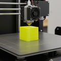 3D printer creating a bright yellow cube on a flat build plate. The printer\'s extruder is Royalty Free Stock Photo