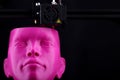 A 3D printer manufactures a humanoid head from pink plastic in dark surrounding - front view, copy space for text