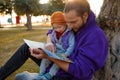 Dad and baby read a book under a tree in the park.