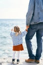 Dad in a denim jacket and jeans holds the hand of a little girl who points to the sea, standing on the stones by the Royalty Free Stock Photo