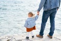 Dad in a denim jacket and jeans holds a little girl by the hand while standing on the stones near the water. Back view Royalty Free Stock Photo