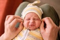 Dad dressing infant daughter. Child is making wry face and unhappy grimace. Hands of father put little striped cap on newborn baby Royalty Free Stock Photo