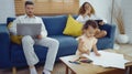 Dad and mom sitting on sofa with computer while daughter painting in living room. Young parent using laptop for works and little