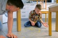 Dad scolds his son for scattered food on the kitchen floor and makes him clean up. Clean up corn flakes off the floor