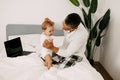 Dad sits on the bed, works on a laptop at home with a baby on his lap, smiles and looks at the screen together Royalty Free Stock Photo