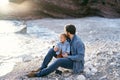 Dad sits on a stone, hugging and kissing the top of the head of a little girl on a pebble beach by the sea against the Royalty Free Stock Photo