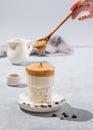 Dalgona coffee. A hand holds a spoon with whipped foam, for instant coffee with ice and milk in a glass on a blue background