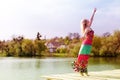 Dancing joy: beautiful blond young slim woman enjoying stretching in long light dress at water lake on summer green outdoors Royalty Free Stock Photo