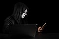 Dangerous anonymous hacker man in black hooded, white mask use computer and mobile phone, break security data corporate server. He Royalty Free Stock Photo