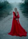Dangerous blonde queen in red fashion lush dress Hides a dagger behind. Backdrop dark fantasy forest in fog. Concept