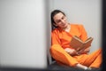 A dangerous mad criminal reads a book sitting on a bunk. In the foreground is the grid of a single cell.