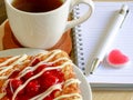 Danish pastry with a cup of hot tea and a pen and small notebook on wood table in morning time and red heart for valentine Royalty Free Stock Photo