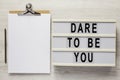 `Dare to be you` on a lightbox, cliboard with blank sheet of paper on a white wooden background, top view. Flat lay, overhead,