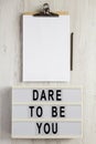 `Dare to be you` on a lightbox, cliboard with blank sheet of paper on a white wooden surface, top view. Flat lay, overhead, from