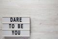 `Dare to be you` on a lightbox, cliboard with blank sheet of paper on a white wooden surface, top view. Flat lay, overhead, from