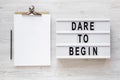 `Dare to begin` words on a lightbox, clipboard with blank sheet of paper on a white wooden background, top view. Overhead, from