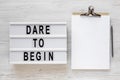 `Dare to begin` words on a lightbox, clipboard with blank sheet of paper on a white wooden surface, top view. Overhead, from abo