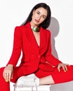 Daring brunette woman in red official pantsuit, jacket with deep neckline on naked body and sneakers sits on a stool Royalty Free Stock Photo