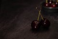 Dark atmospheric image of two ripe red cherries with crossed stems and a ceramic bowl full of cherries in the background on a dark