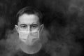 Dark black and white image. Caucasian adult male in protective mask surrounded by smoke.