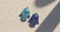Two hamsa hand charms are positioned on sand. One is turquoise, and the other is Royalty Free Stock Photo