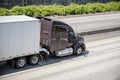 Dark brown big rig commercial long haul semi truck transporting dry van semi trailer on the divided highway road