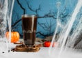 Dark brown coffee in tall glass mug on hot plate with coffee beans, cinnamon sticks, spider, pumpkins and cobwebs against night