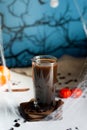 Dark brown coffee in tall glass mug on hot plate with coffee beans, cinnamon sticks, spider, pumpkins and cobwebs against night